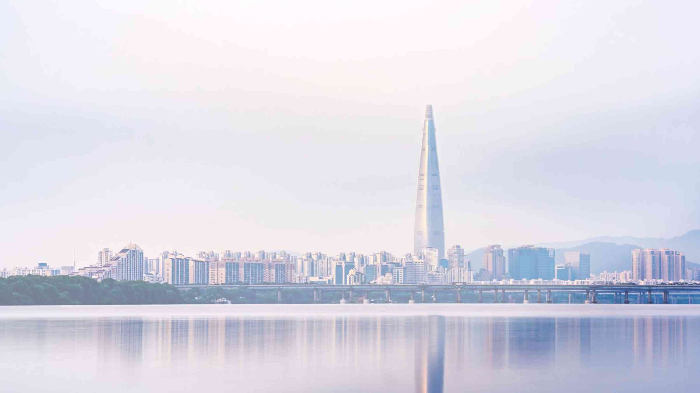 The skyline of a large city with a modern skyscraper soaring above the other buildings, reflected in the water. The photo has a purple hue and was likely taken at dusk.